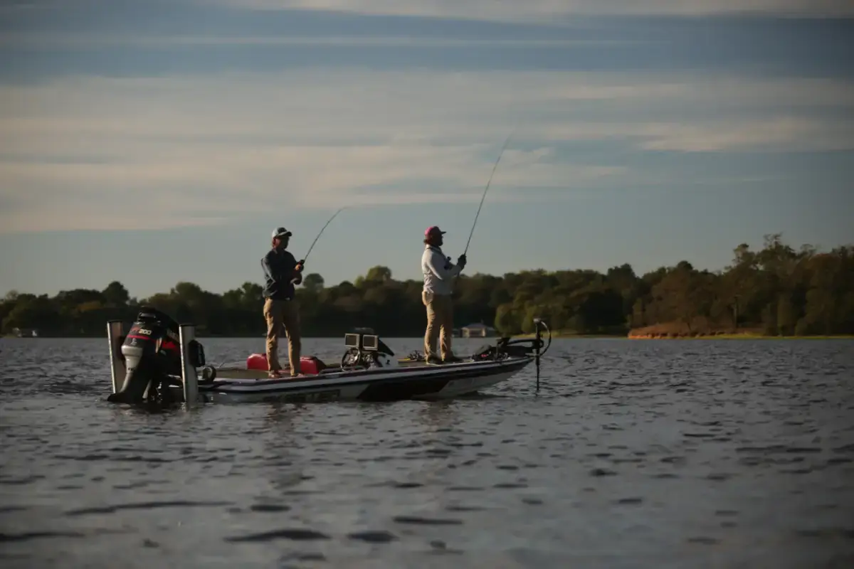 Two anglers casting from a fishing boat on calm water at dusk, a wooded shoreline behind them