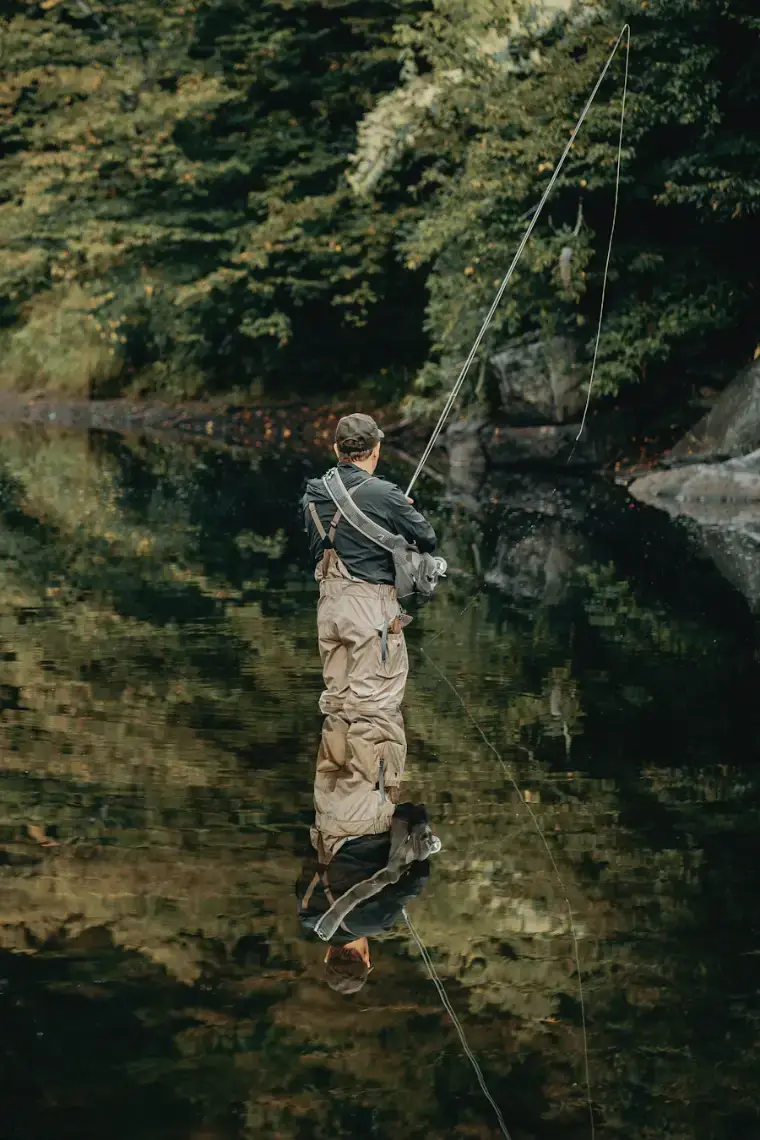 An angler in waders casting a fly rod, reflected in still green water