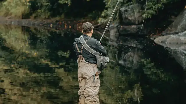 An angler in waders casting a fly rod, reflected in still green water