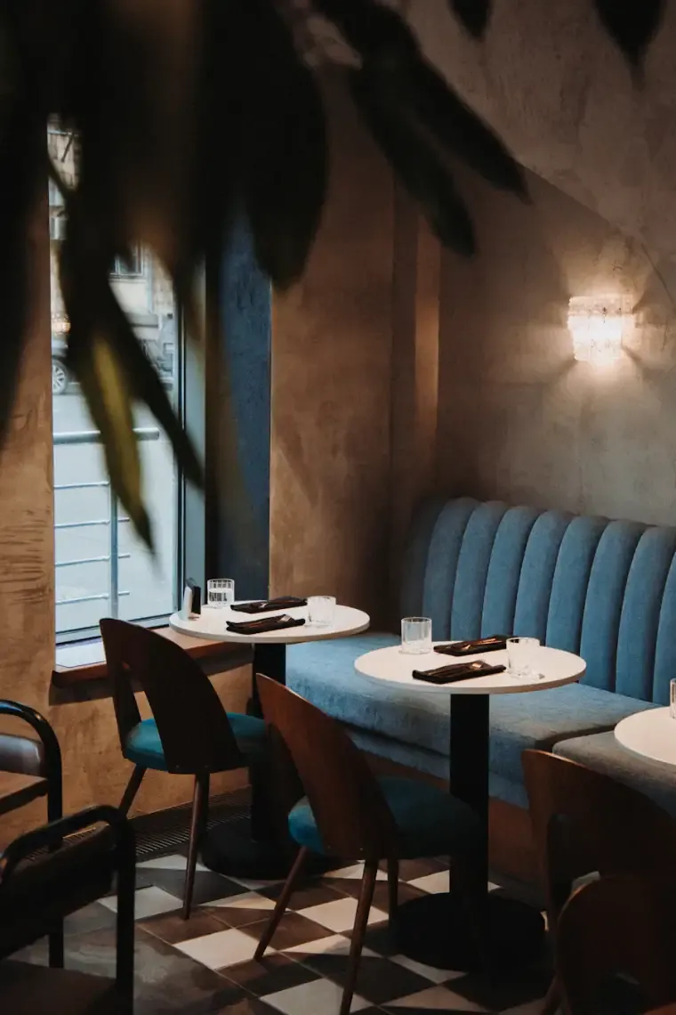 A quiet dining room with teal velvet booths, round tables set for supper and a lamp glowing on the wall