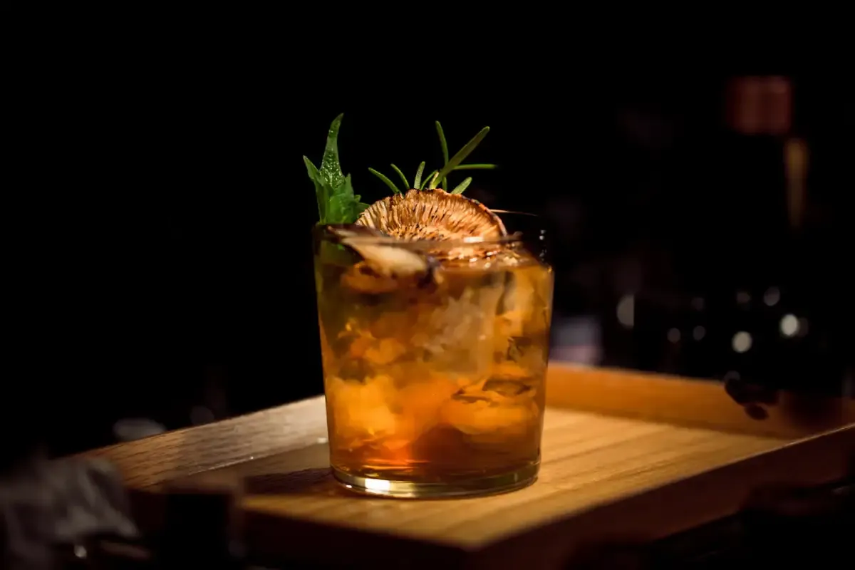 An old fashioned with a dried orange wheel and rosemary on a wooden bar tray in low light