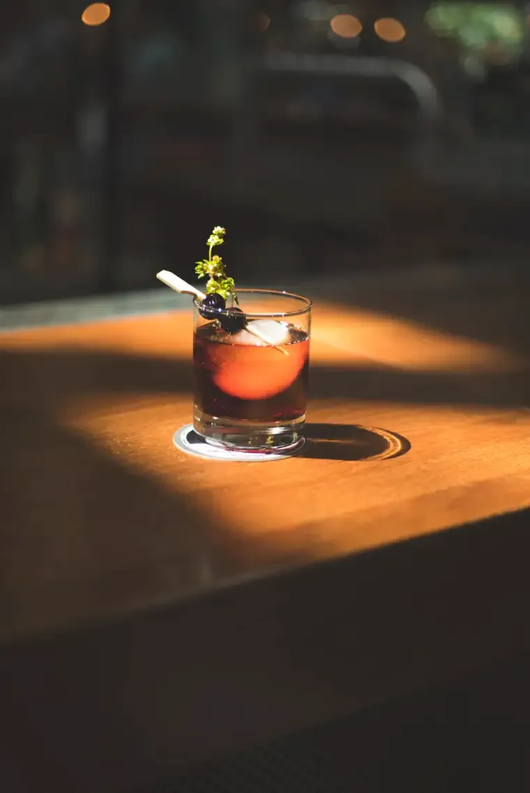A brandy old fashioned with an orange slice and herb sprig, lit by a single lamp on the dark wood bar