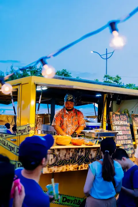 A cook hands out food from a yellow food truck under strings of lights at dusk