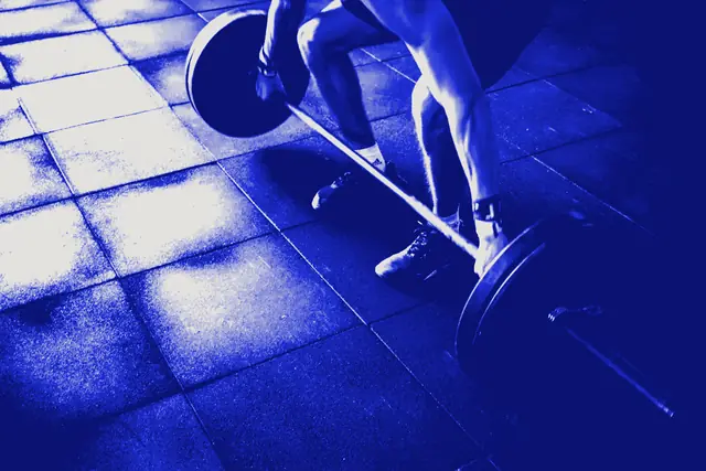 A lifter in trainers pulling a loaded barbell off rubber gym flooring.