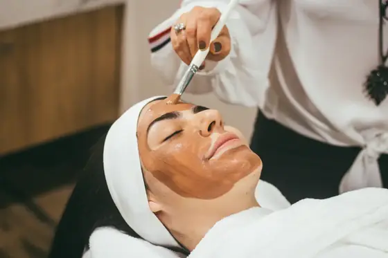 An esthetician brushing a clay mask onto a client’s face, her hair wrapped in a white towel