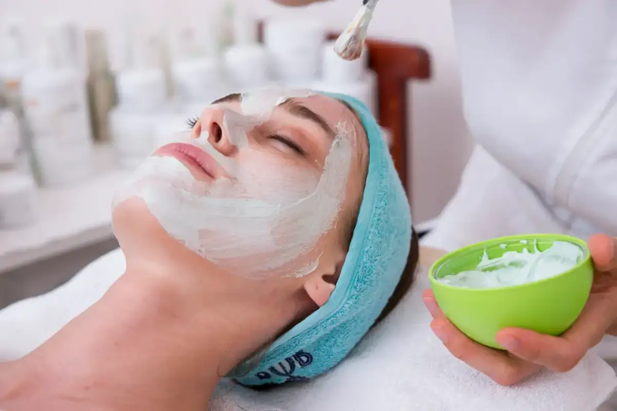A client lying back with a white cream mask on her face while a provider holds a small bowl of it
