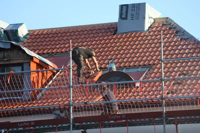 A roofer kneeling on a red tile roof behind scaffolding and safety netting